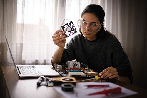 Photo of a student at home in front of a computer, wearing safety googles and examining a piece of computer hardware.