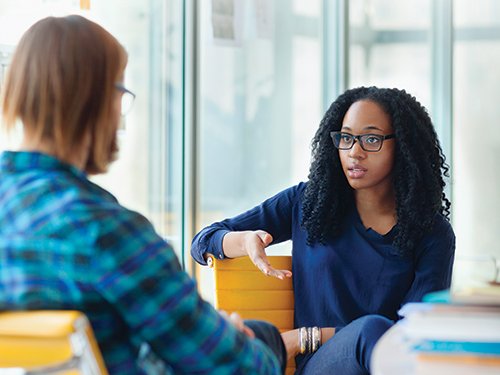 A White and Black woman are sitting at a desk having a discussion.