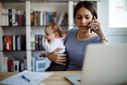 Photo of a woman sitting in front of a laptop holding a young child while on the phone