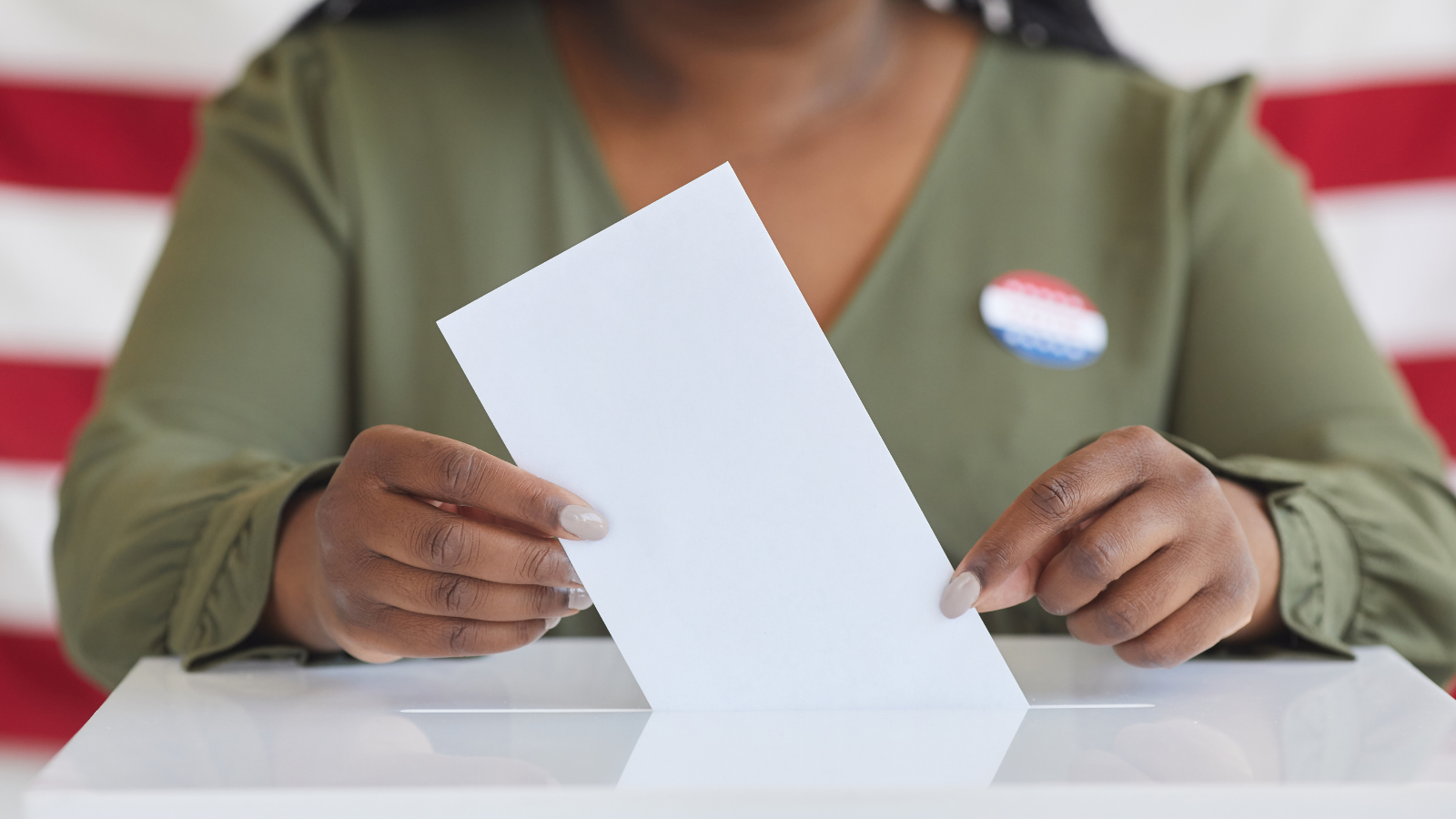 Photo of a Black woman pictured from neck down casting her vote in a ballot box while posed in front of American flag.