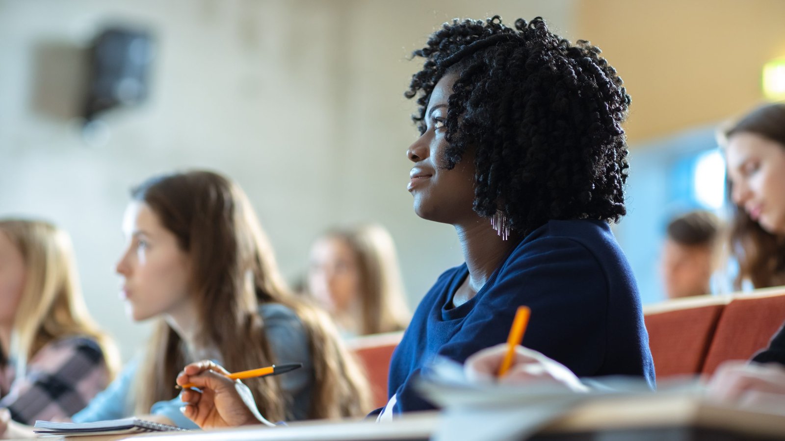 Photo of a Black woman student sitting in a lecture hall surrounded by peers.