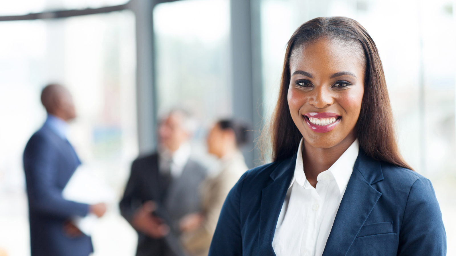 Photo of a Black woman in business attire standing in the foreground with other people in the background