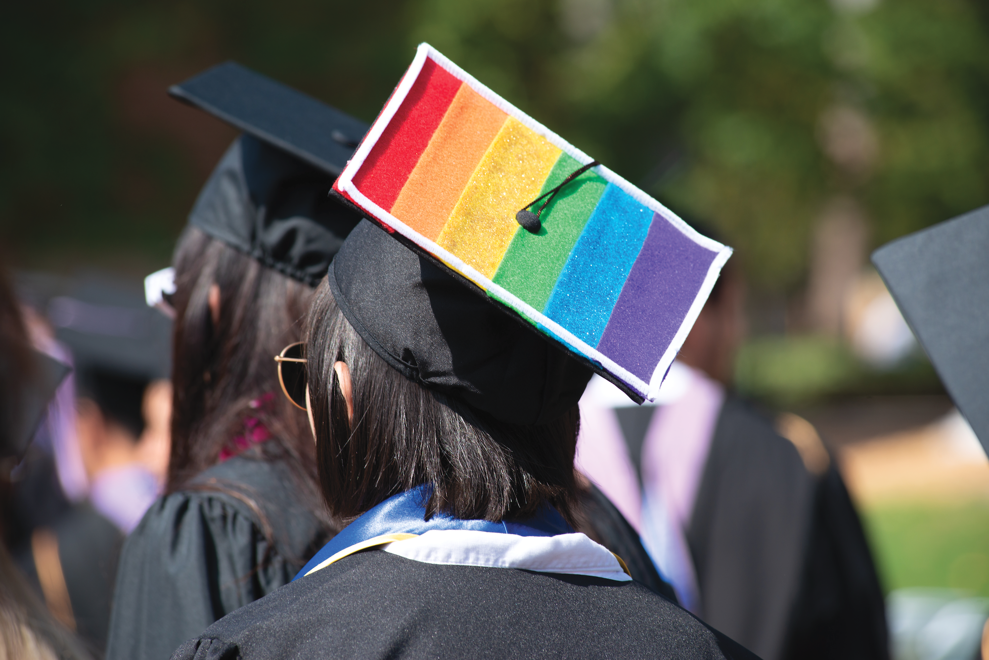 Photo of a student turned away from the camera wearing a graduation cap that is painted in the colors of the LGBTQ Pride flag